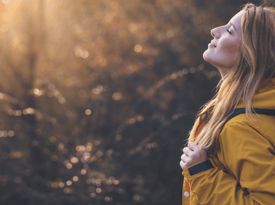 Person standing outdoors with eyes closed, breathing in soft sunlight through trees, symbolising mindfulness, reflection, and emotional awareness.