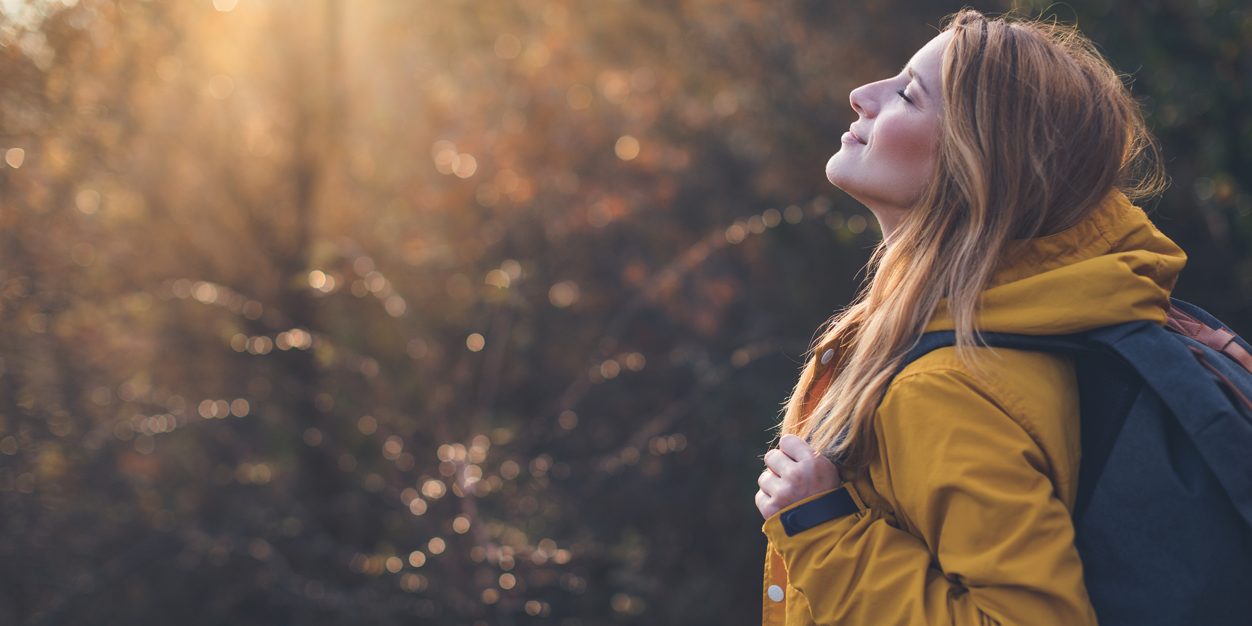 Person standing outdoors with eyes closed, breathing in soft sunlight through trees, symbolising mindfulness, reflection, and emotional awareness.