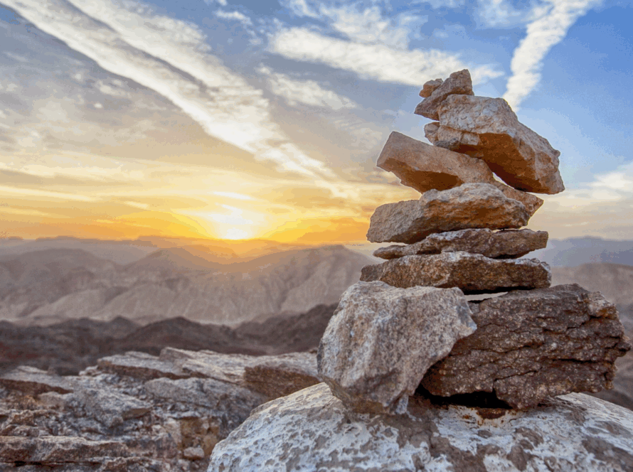Stacked stones on a mountain peak at sunrise with soft clouds and wide desert hills in the background.