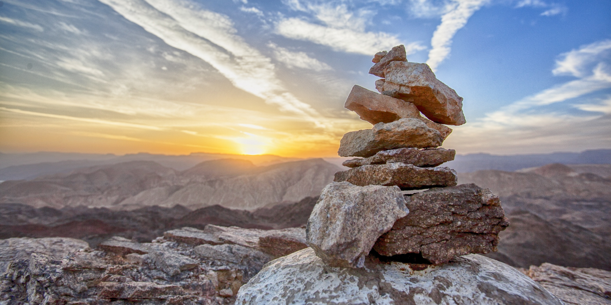 Stacked stones on a mountain peak at sunrise with soft clouds and wide desert hills in the background.